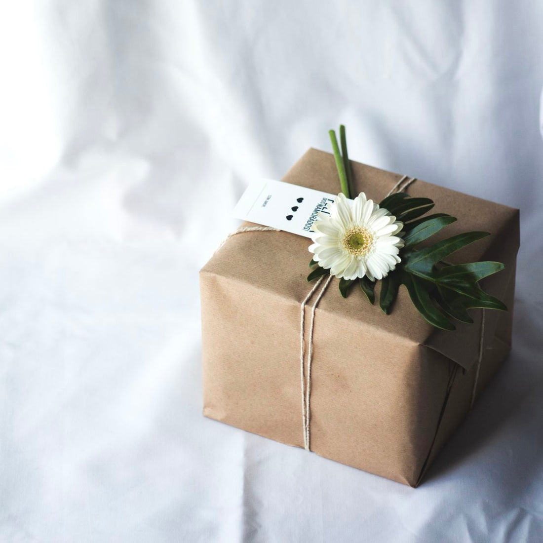 A brown paper wrapped present with a white ribbon tied around it. A daisy is placed on top of the ribbon for decoration.