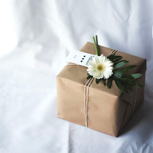 A brown paper wrapped present with a white ribbon tied around it. A daisy is placed on top of the ribbon for decoration.